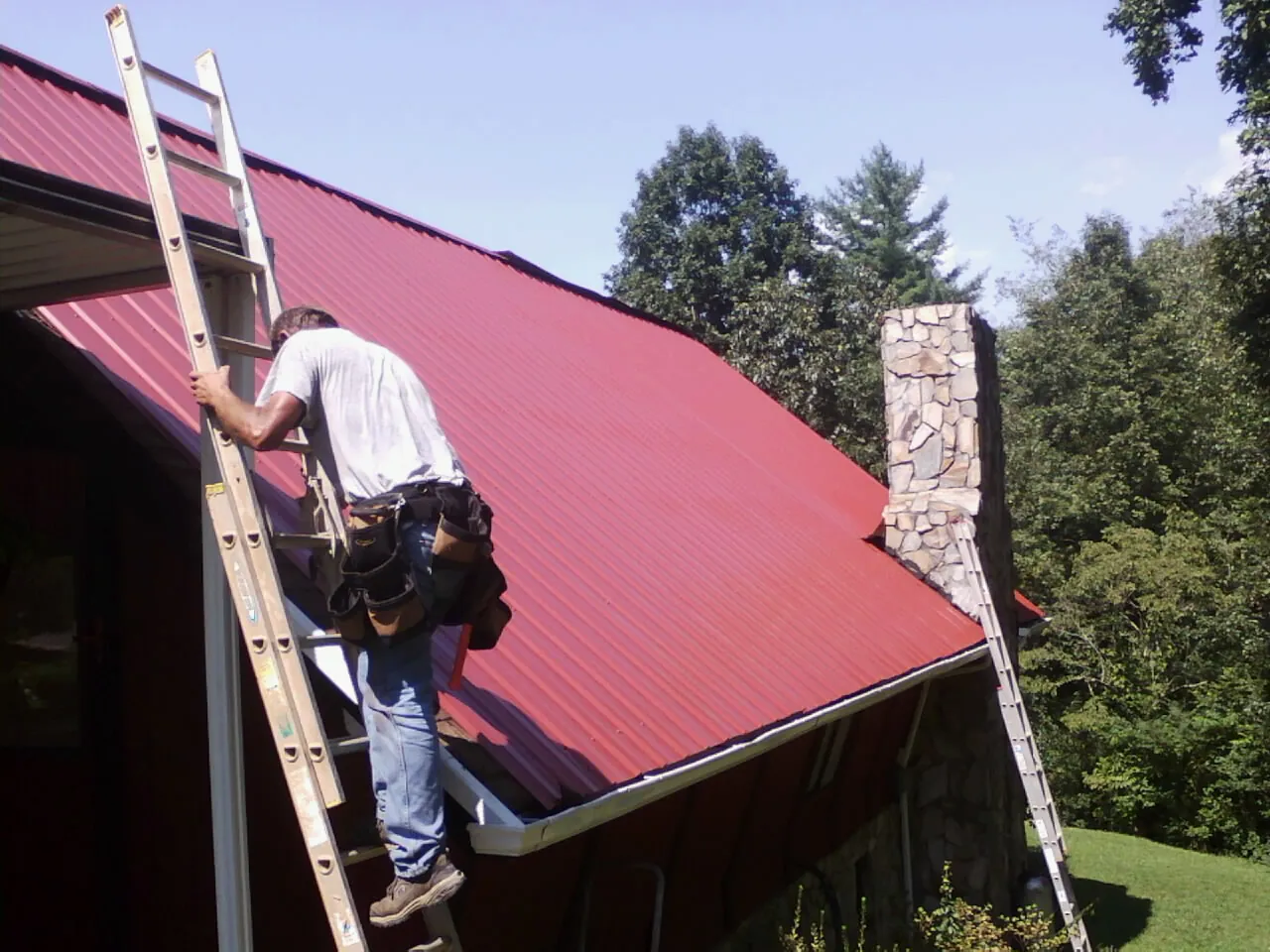 Roofer on a ladder beside a red metal roof and stone chimney — Soffit & Fascia Repair by Roofing Brothers in Woodbury Heights